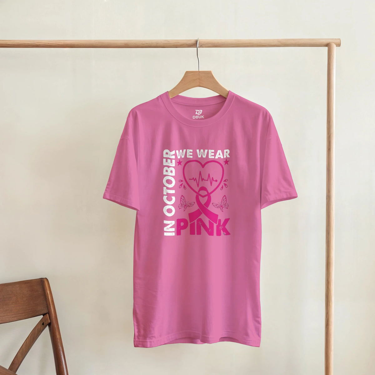 Pink unisex T-shirt hanging with 'OCTOBER WE WEAR PINK' heart ribbon graphic