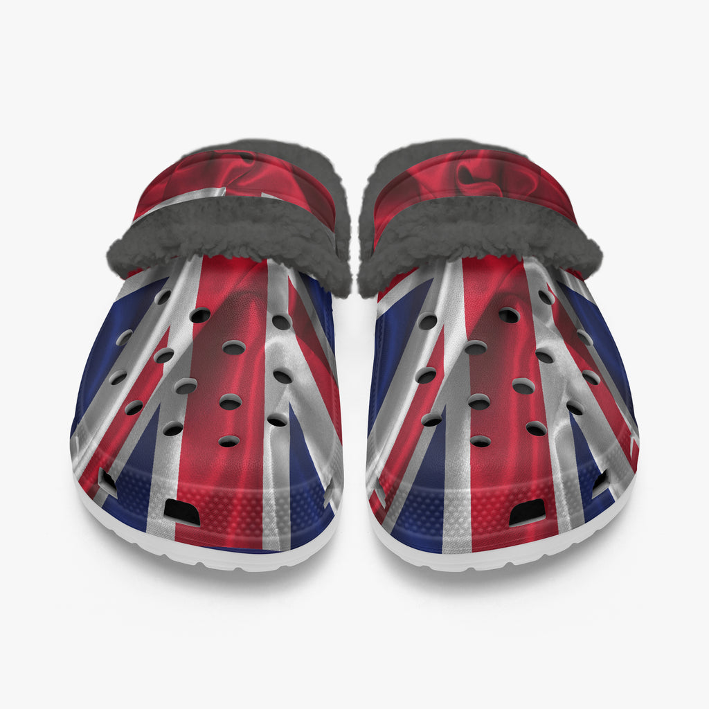 A pair of clog-style shoes with a Union Jack flag print in red, white, and blue, featuring a gray faux-fur trim along the top strap, shown against a light gray background.