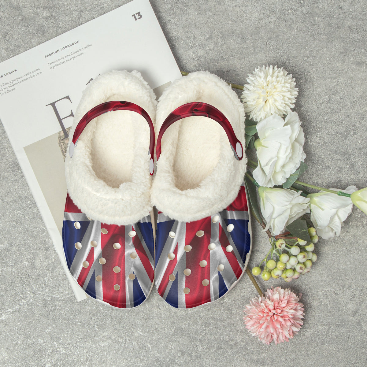 A pair of plush slipper-style shoes with white sherpa-like interiors and red trim, decorated with Union Jack flag patterns and silver studs, arranged on a gray surface with a magazine and white flowers.