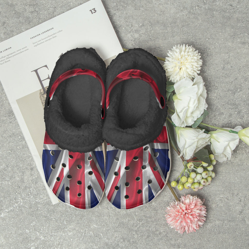 A pair of black fuzzy slip-on shoes with red velvet straps and Union Jack flag footbeds, styled on a gray stone surface with white flowers and a magazine.