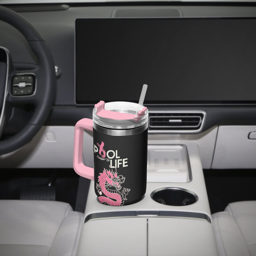 A black insulated travel tumbler with a pink handle, lid, and straw sits in the center console of a car. The tumbler has a pink dragon graphic and the words 'POOL of LIFE' printed on it.